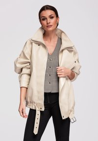 Femme portant une veste en cuir beige surdimensionnée par-dessus un gilet à carreaux boutonné et un pantalon noir, posant devant un fond uni
