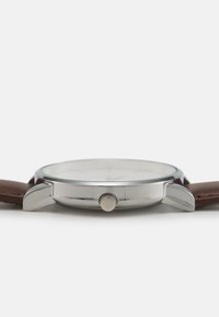 Montre en acier inoxydable avec un cadran en verre transparent, un bracelet en cuir marron, un boîtier rond et une couronne argentée texturée sur le côté.