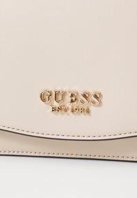 Borsa a tracolla sintetica beige pallido con logo dorato "GUESS EST 1981", design con patta curva e dettagli di cucitura a contrasto.