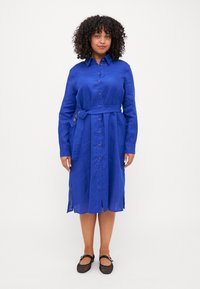 Vestito camicia in lino blu con maniche lunghe, chiusura a bottoni frontale, vita con cintura e spacchi laterali. Indossato con scarpe in rete nere.