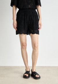 Shorts en œillet noirs avec des bords festonnés, une taille élastique et de subtils motifs découpés. Associés à des sandales noires à larges brides.