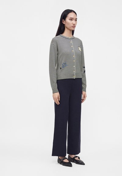 Femme portant un cardigan rayé noir et blanc boutonné avec broderie, un pantalon large bleu marine et des ballerines noires pointues, debout de profil.