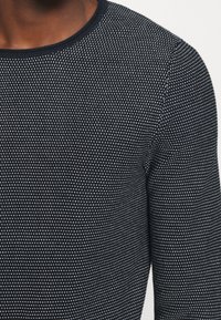 Chemise à manches longues bleu marine avec un motif texturé à pois et un col rond. Le tissu semble doux et léger.