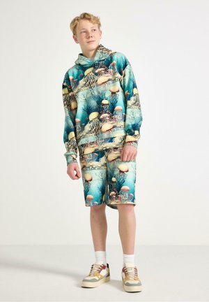 Teenagedreng iført matchende blå-grøn hættetrøje og shorts med blæksprutteprint, stående med hænderne afslappede, kigger let opad og til siden.