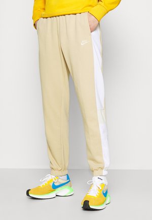 Person iført beige og hvide Nike træningsbukser med gule, blå og grønne Nike sneakers og en gul sweatshirt, stående med hænderne i lommerne.