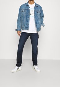 Džínová bunda se dvěma náprsními kapsami, bílý tričko s grafickým potiskem v pestrých barvách, tmavě modré džíny a bílé tenisky s černými akcenty.