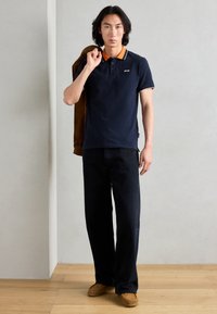 Mėlynas polo marškinėliai su oranžiniais apykaklės akcentais ir baltais krašteliais, derinami su tamsiomis džinsomis ir ruda avalyne. Modelis laiko rudą švarką.