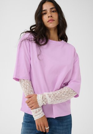 Femme aux longs cheveux foncés portant une chemise lilas à manches courtes sur des manches longues en dentelle blanche, un jean bleu, et une bague et un bracelet en argent.