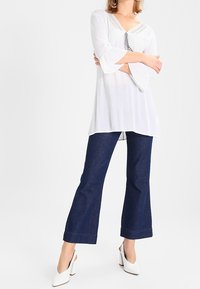 Tunic bianca con maniche a campana, tessuto testurizzato e dettagli in pizzo nella scollatura, abbinata a jeans a zampa d'elefante blu scuro e tacchi bianchi.
