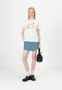 Biely bavlnený tričko s farebnými potlačami, džínsová sukňa, čierne topánky a čierna taška. Model má na sebe biele ponožky. Jednoduché pozadie.