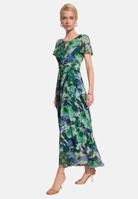 Maxi sukienka z przezroczystej tkaniny, zdobiona zielonym i fioletowym wzorem kwiatowym. Krótkie rękawy, dopasowana talia i zwiewna spódnica. Widać sandały na obcasie.