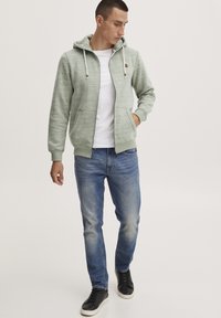Veste à capuche zippée vert clair avec cordons, poches avant, accents gris, associée à un jean bleu et des baskets noires. T-shirt blanc uni en dessous.