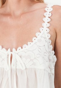Witte kanttop met bloemenpatronen, verstelbare strik aan de voorkant en geschulpte kantbandjes; lichtgewicht stof met een zachte textuur.