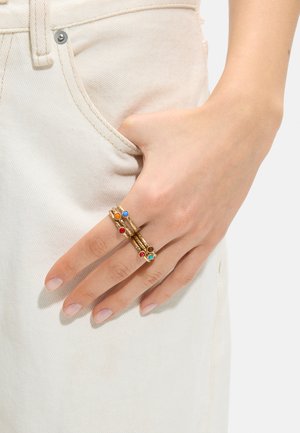 Ręka z pierścionkami z kolorowymi kamieniami szlachetnymi spoczywa w kieszeni jasnobeżowych dżinsów.