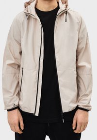 Ljusbeige vindjacka med huva, svart dragkedja och mudd. Har sidofickor och en avslappnad passform. Bärs över en svart tröja.