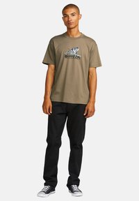 Khaki cotton t-shirt with a graphic design featuring a butterfly and text on the front, paired with black pants and black sneakers.