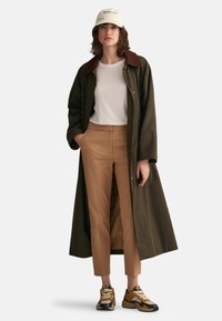 Long olive green coat with a brown collar, white shirt, tan trousers, and multicolored sneakers. Person has hands in pockets.