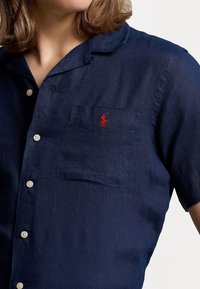 Navy blue short-sleeve shirt made of linen, featuring a pocket with an embroidered red polo logo and white buttons along the front.