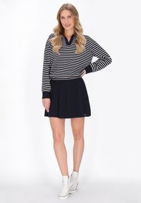 Navy and white striped pullover, rentoitu istuvuus V-pääntiellä; pleidattu navy-hame; valkoiset korkeat tennarit värikkäillä korostuksilla.