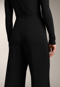 Person iført tætsiddende sort langærmet top og højtaljede, vidde sorte bukser, stående med den ene hånd afslappet ned langs siden.