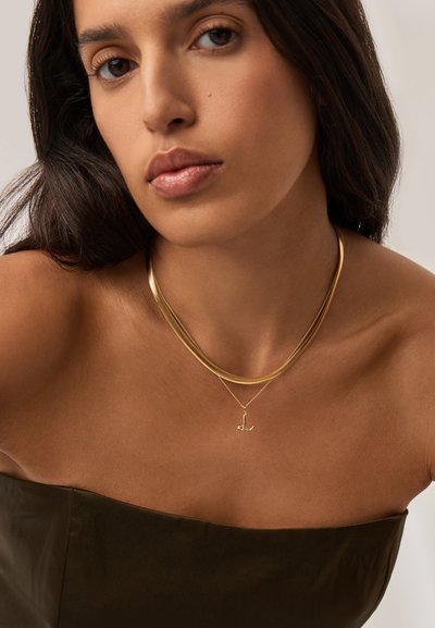 Γυναίκα με σκούρα μαλλιά φοράει πολυεπίπεδα χρυσά κολιέ, συμπεριλαμβανομένου ενός με μενταγιόν με το γράμμα "L", και ένα στράπλες τοπ σε σκούρο ελαιδί χρώμα.