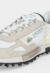 Λευκό και μπεζ sneaker Lacoste με πράσινο λογότυπο, λευκά κορδόνια, υφασμένη σόλα και τυπωμένες γεωγραφικές συντεταγμένες στο πλάι.