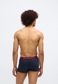 Muškarac s kovrčavom kosom prikazan s leđa, nosi mornarsko plave bokserice s tamnocrvenim pojasom, stoji naslonjen na jednoličnu svijetlu pozadinu.