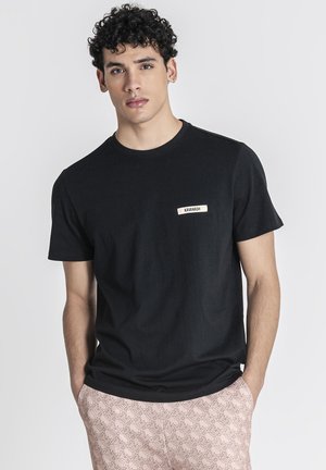 Giovane uomo con capelli ricci che indossa una maglietta nera con etichetta Kavanagh e pantaloni beige a fantasia, in piedi con le mani in tasca.