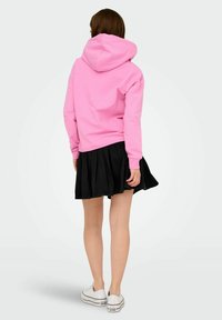 Rosa huvtröja med stor huva, matchad med en svart veckad kjol och vita sneakers. Texturerna är mjuka och avslappnade.