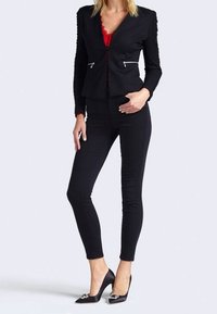 Blazer noir ajusté avec des manches texturées, sous-vêtement en dentelle rouge et accents zippés, associé à un pantalon skinny noir et des talons noirs.