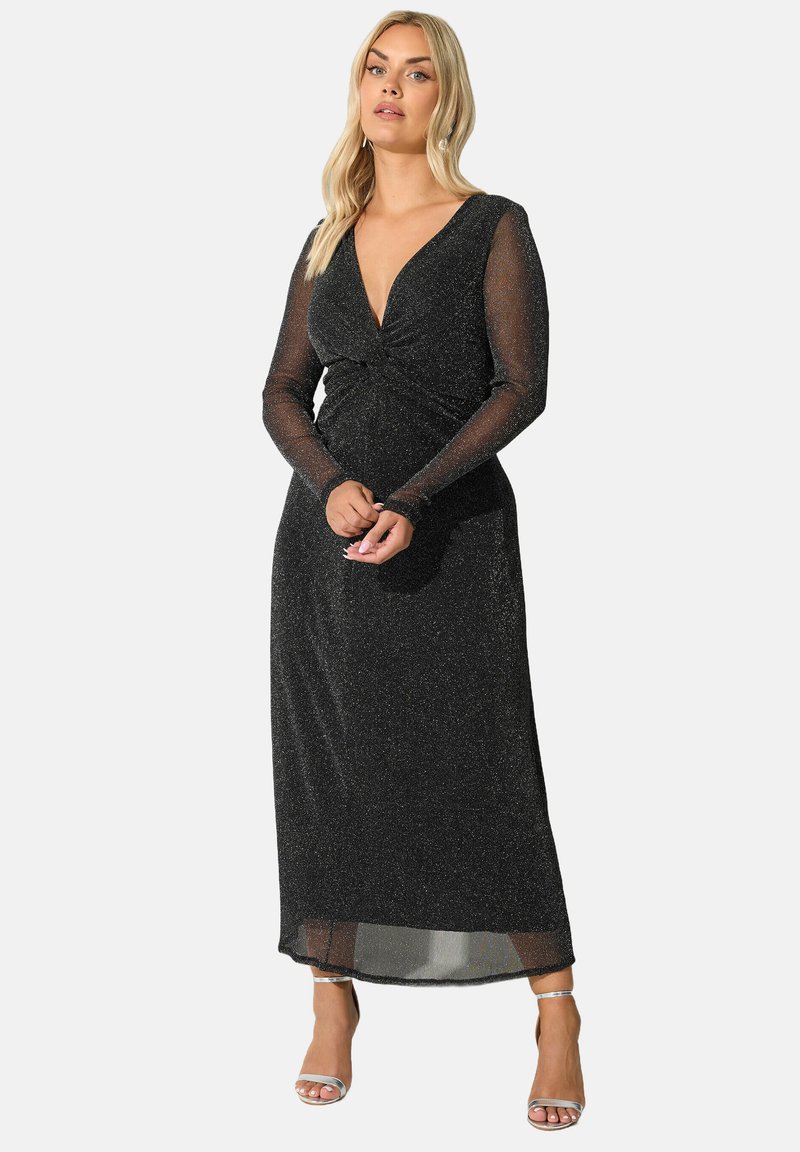 Pitkä musta mekko, jossa on syvä V-pääntie ja pitkät läpikuultavat hihat. Kankaassa on hienovarainen kiilto. Solmutyylinen yksityiskohta vyötäröllä, nilkkoihin ulottuva.