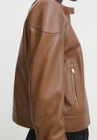 Brun läderjacka med dragkedjeficka på sidan, bärs över svarta byxor, visad från sidan mot en enfärgad bakgrund.