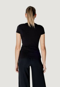 Maglietta nera a maniche corte, design aderente con texture liscia. Chi la indossa ha lunghi capelli raccolti in una coda di cavallo, abbinati a pantaloni neri a gamba larga.