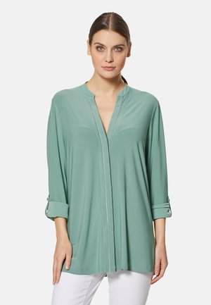 Mintgroene blouse met lange mouwen, gemaakt van zachte, lichte stof, met een V-hals met zilveren rand en opgerolde mouwen.