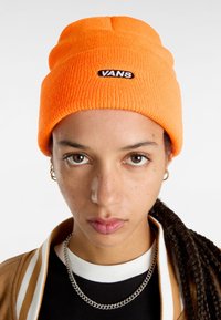 Gorro de punto naranja con vuelta, que presenta un logo negro de "VANS". El modelo lleva una chaqueta marrón con una camiseta de rayas blanca y negras debajo.