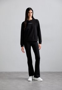 Μαύρο φούτερ με λαιμόκοψη και λευκό κείμενο, συνδυασμένο με μαύρα φουσκωτά παντελόνια και λευκά αθλητικά παπούτσια, φωτογραφημένο σε ουδέτερο φόντο.