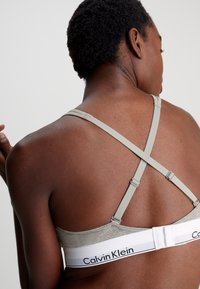 Grå bh med en glatt tekstur, som har krysset stropper og et hvitt elastisk bånd merket "Calvin Klein" bak.
