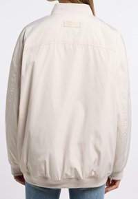 Persona che indossa una giacca leggera beige chiaro, vista da dietro, abbinata a jeans blu su uno sfondo bianco uniforme.