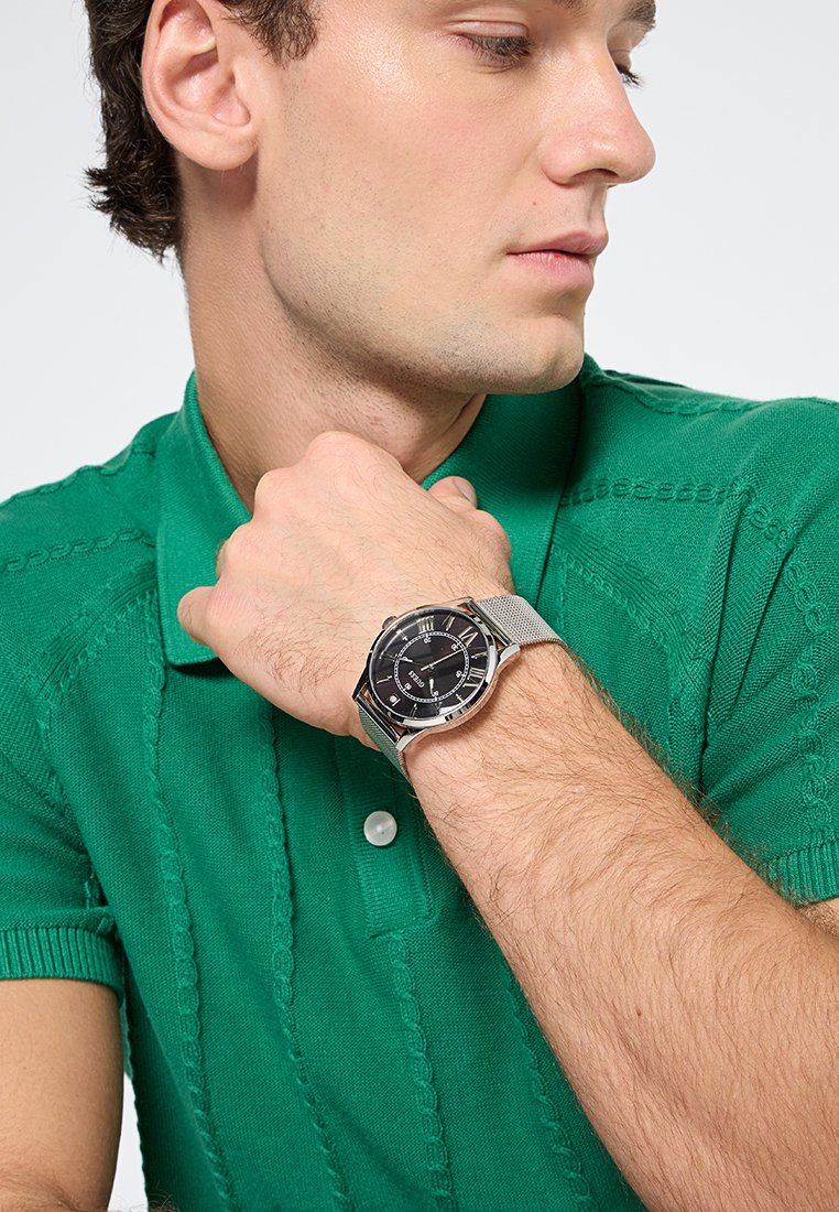 Reloj con correa de malla plateada, esfera negra, números romanos y caja redonda de acero inoxidable, llevado en una muñeca sobre una camisa polo verde.