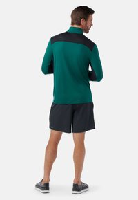 Långärmad teal idrottströja med svarta detaljer, kombinerad med svarta shorts och mönstrade sneakers, sedd bakifrån.