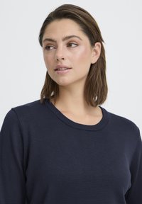 Pull-over en maille bleu marine avec encolure dégagée. Présente une texture lisse et des manches longues, ajusté près du corps.