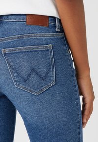 Jeans de denim azul con cintura alta, que cuentan con un parche de cuero marrón etiquetado "Wrangler", y un diseño distintivo de bolsillo trasero cosido.