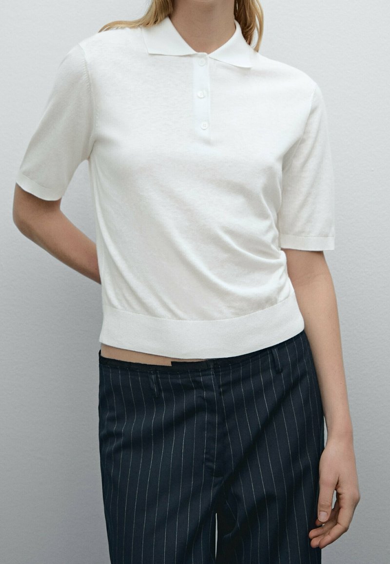 Personne portant un polo blanc à manches courtes et un pantalon noir à fines rayures, se tenant avec une main derrière le dos.