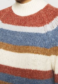 Sweter w paski w kolorach czerwonym, kremowym, niebieskim i brązowym. Dzianinowa tekstura z wysokim kołnierzem i luźnym krojem.