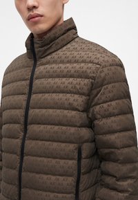 Homme portant une doudoune marron avec un motif répété du logo "AX" et une fermeture éclair noire sur fond blanc uni.