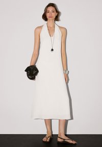 Weißes Trägerkleid mit Halsband, knöchellang, aus leichtem Material. Kombiniert mit einer schwarzen geflochtenen Clutch und braunen Sandalen mit Riemchen.