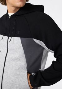 Mann in schwarzem, dunkelgrauem und hellgrauem Reißverschluss-Hoodie mit Kapuze, mit der Hand in der seitlichen Tasche, gezeigt von den Schultern bis zur Taille.