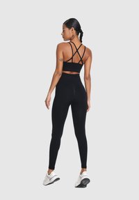 Žena s pletenými vlasmi, oblečená v priliehavých čiernych legínach, čiernom športovom podprsenku s remienkami a bielych športových topánkach, stojaca pred čistým pozadím.