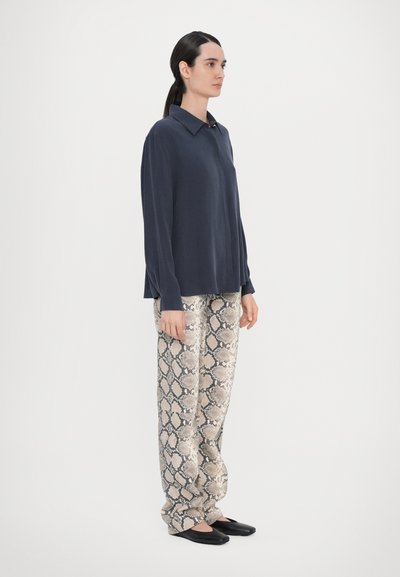 Cămașă albastră cu mânecă lungă și guler, asortată cu pantaloni bej cu imprimeu de șarpe. Modelul poartă pantofi negri, fiind așezat cu spatele la un fundal alb.