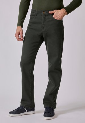 Homme portant un pantalon droit vert foncé, une chemise verte à manches longues et des baskets noires à semelles blanches, debout avec une main dans la poche.